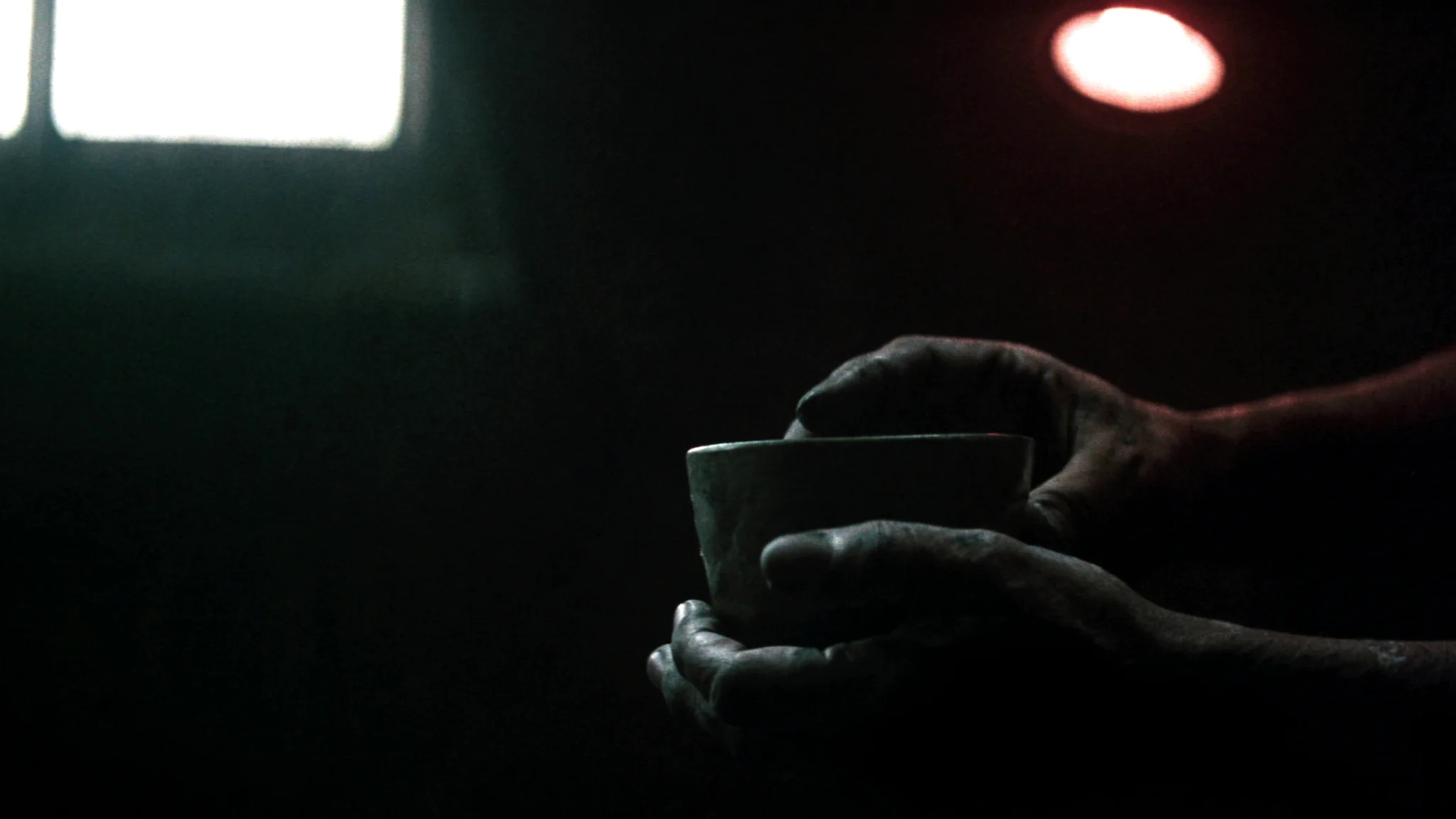 A finished stoneware cup held in two clay-dusted hands, lit by a warm lamp.